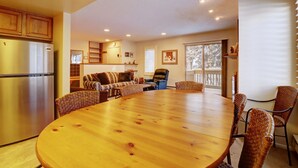 A spacious living area with a large wooden dining table surrounded by wicker chairs, a kitchen with a stainless steel refrigerator, and a cozy seating area with a sofa and armchairs.