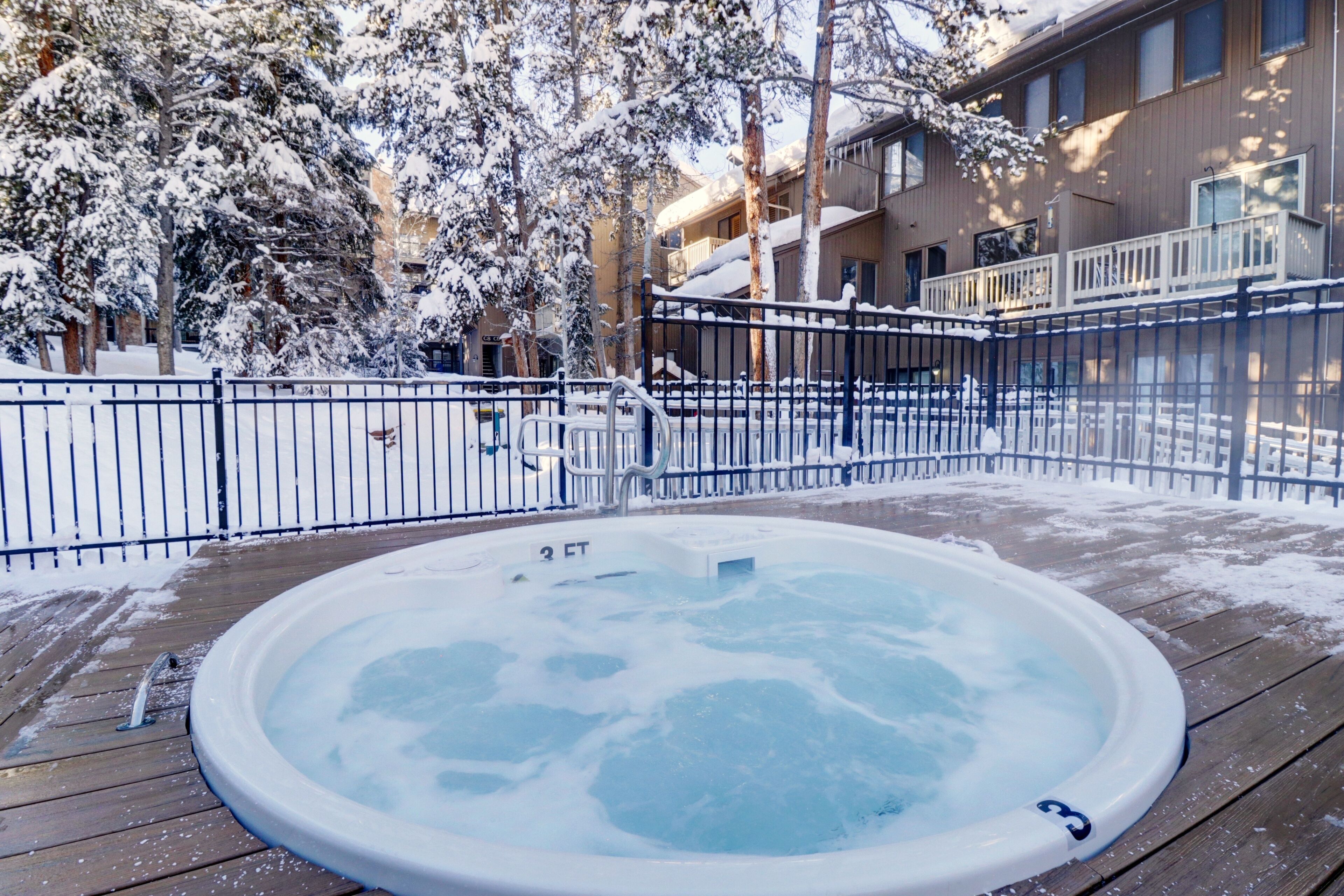 A hot tub on a wooden deck surrounded by snow-covered trees and a fence.