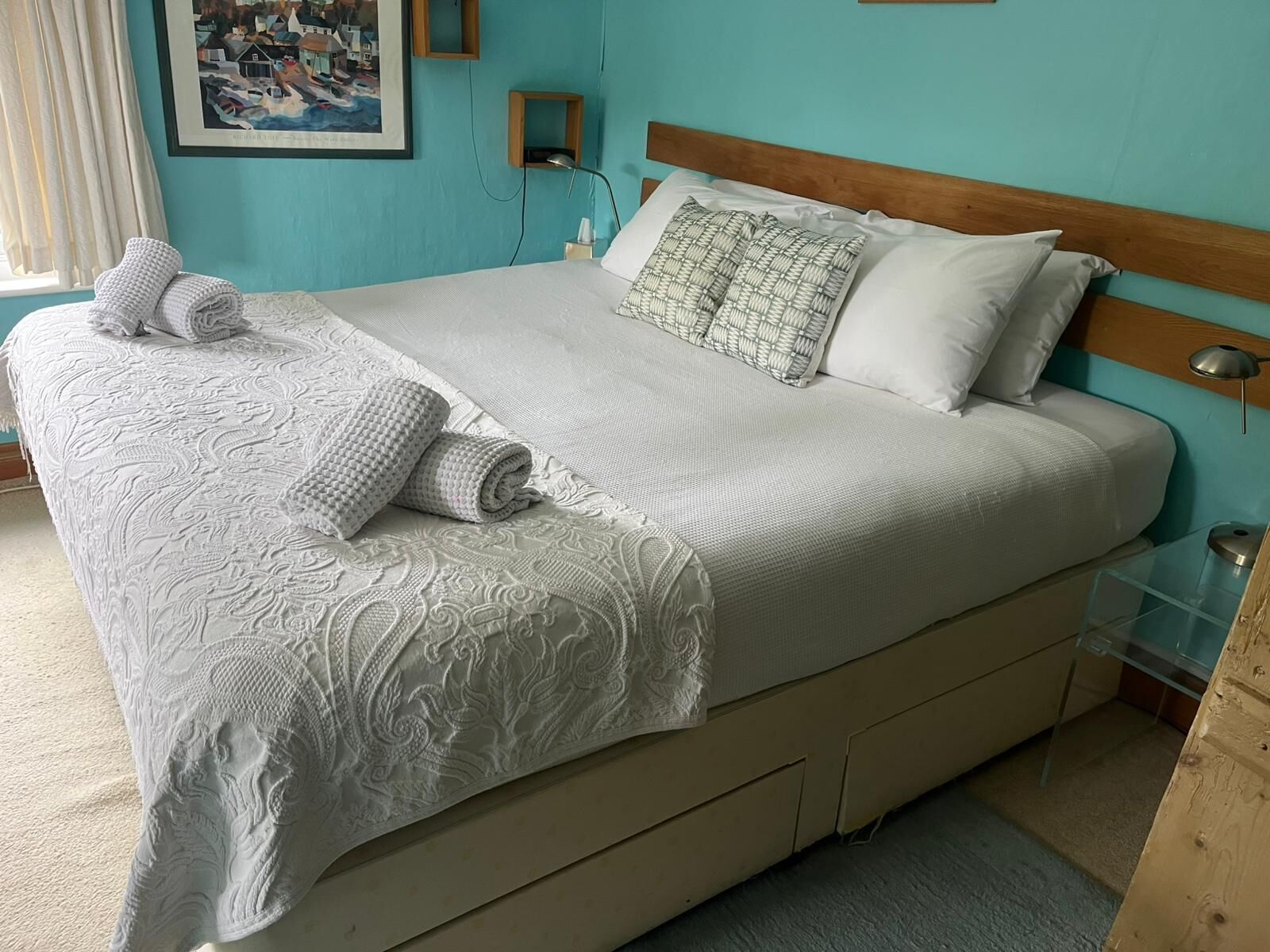 Una cama bien hecha con una colcha blanca y almohadas, un cabecero de madera y una mesita de noche con una lámpara.