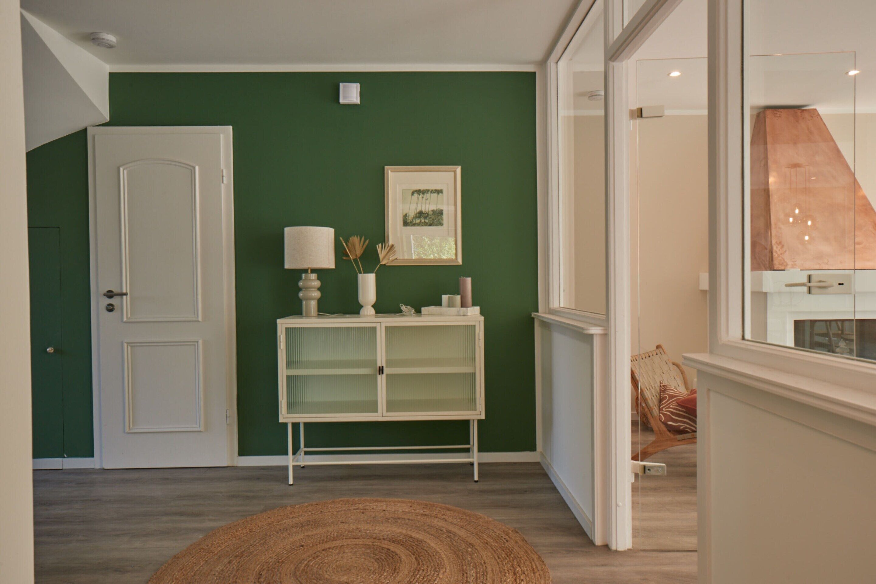 Een gang met een groene muur, een witte deur, een witte console met een lamp en een lijst met een afbeelding.