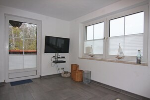 A room with a television, a window with blinds, a small shelf with decorative items, and a wicker basket.