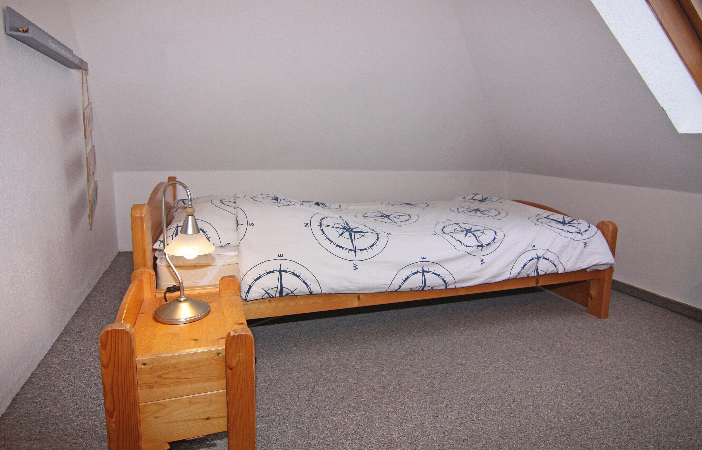 Una stanza piccola con un letto singolo, una struttura in legno, una scrivania con una lampada e una coperta con motivo a bussola.