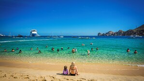 A beach with clear turquoise water, people swimming and sunbathing, and a cruise ship in the background.