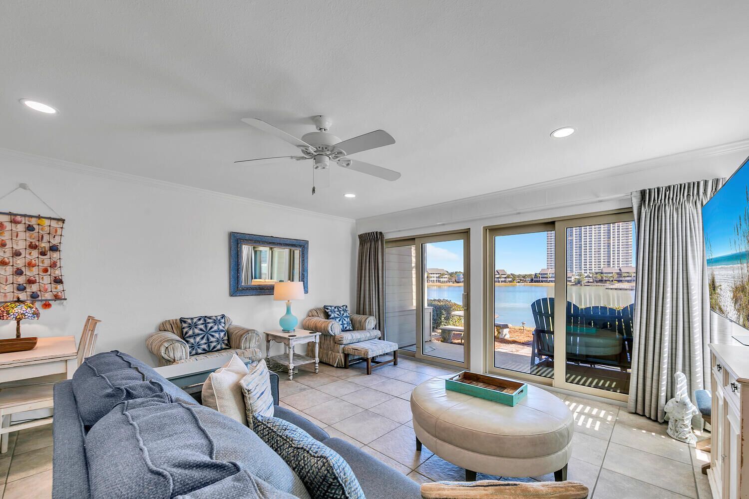 A spacious living room with a ceiling fan, a large window with a view of the beach, and a dining area with a table and chairs.