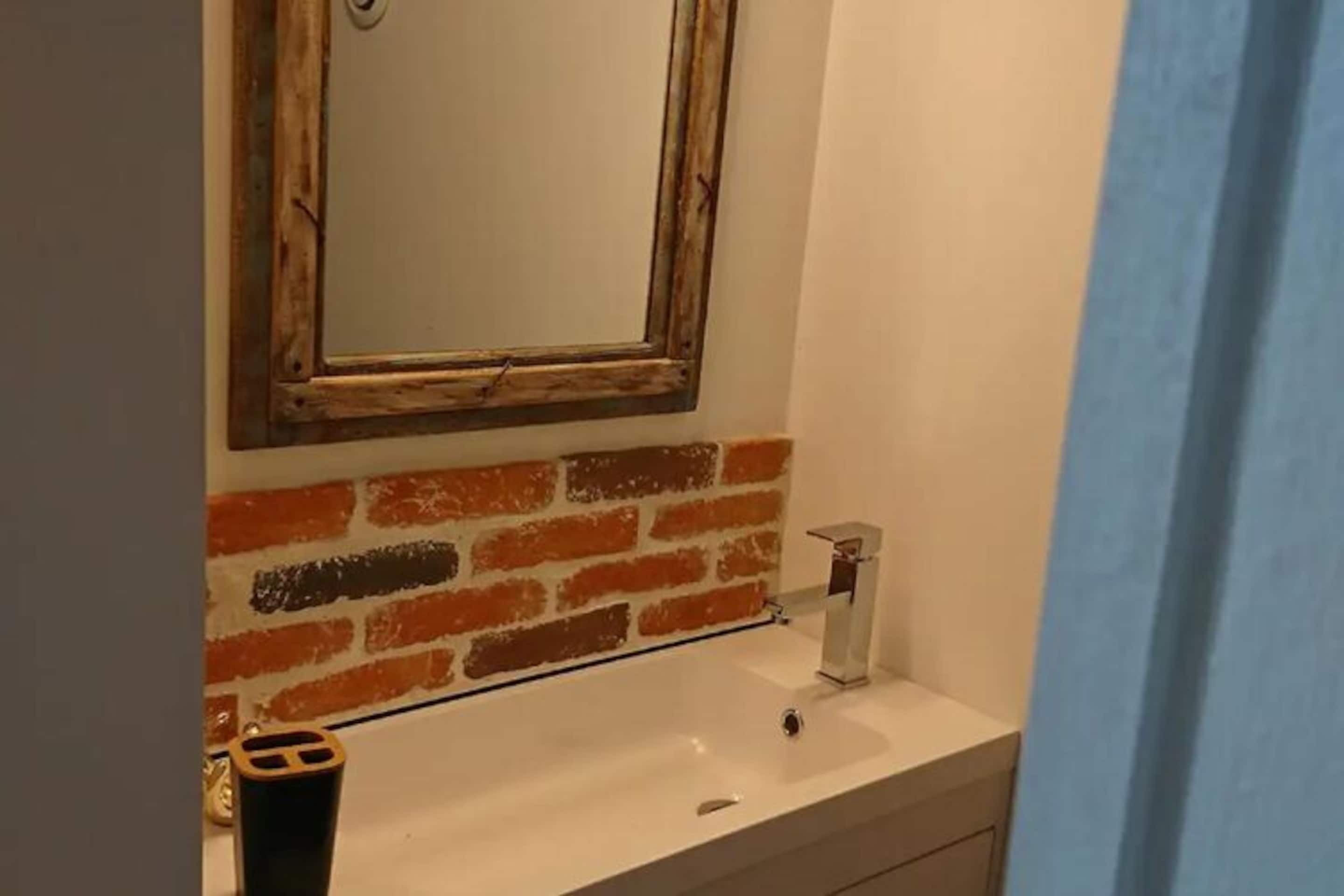 Un baño con lavamanos blanco, un espejo con marco rústico y una pared de ladrillo.
