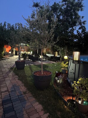 Un espacio al aire libre pavimentado con plantas en macetas, una zona de estar con sillas y mesas, y una piscina con lámpara.