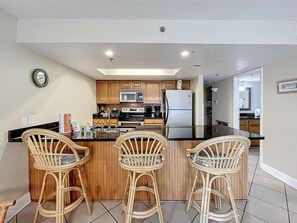 A kitchen with a central island, wooden cabinets, a refrigerator, and a microwave. There are three wicker chairs around the island.