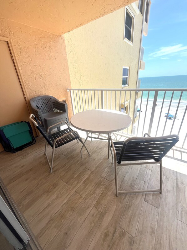 Un balcón con una mesa redonda y dos sillas, una maleta verde, y vistas a la playa y al océano.