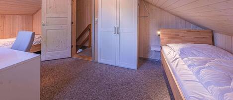 A bedroom with a sloped ceiling, wooden walls, a bed, a desk, and a wardrobe.