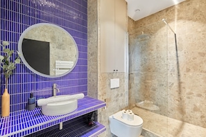 A bathroom with a blue tiled wall, a round mirror, a sink, a toilet, and a glass shower enclosure. - Charming Apartment Center of Paris (Paris)