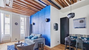 Un soggiorno moderno con una parete blu a contrasto, travi in legno a soffitto, un divano con cuscini blu, un'area pranzo con sedie e un tavolo, e una lampada a sospensione.