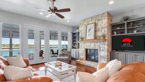 A spacious living room with a stone fireplace, a ceiling fan, and a large TV screen displaying a logo. - Lakeside Dreamhouse I Lake, Hot tub, Dogs (Canyon Lake)