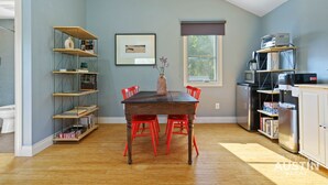 A compact kitchen with a dining table, red chairs, a bookshelf, and a window with a view.