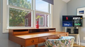 A room with a wooden desk, a window with a view of a red building, a cactus-patterned chair, and a TV displaying streaming services.