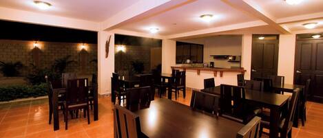 A spacious dining area with multiple tables and chairs, a kitchen in the background, and a brick wall with plants.