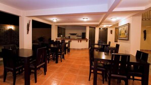 A spacious dining area with multiple tables and chairs, a kitchen in the background, and a stone wall on one side. - Hotel El Cerro (Liberia)