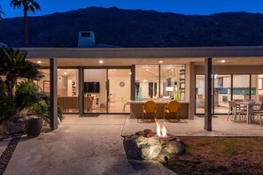 Une maison moderne dotée d’un patio comprenant un foyer extérieur, des sièges et une cuisine visible à travers des portes vitrées.