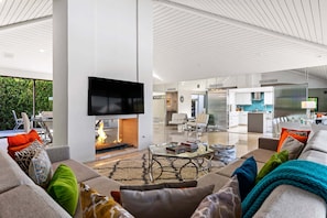 A modern living room with a fireplace, a flat-screen TV, and a dining area with a kitchen in the background.