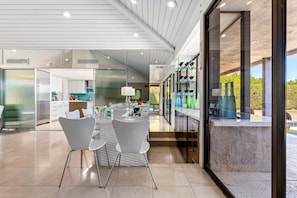 A modern kitchen with a central island, white chairs, and a dining area with a view of greenery.