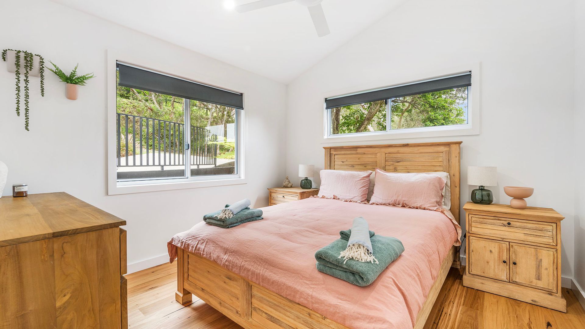 A bedroom with a wooden bed, two bedside tables, a nightstand, a desk, and a window with a view of greenery.
