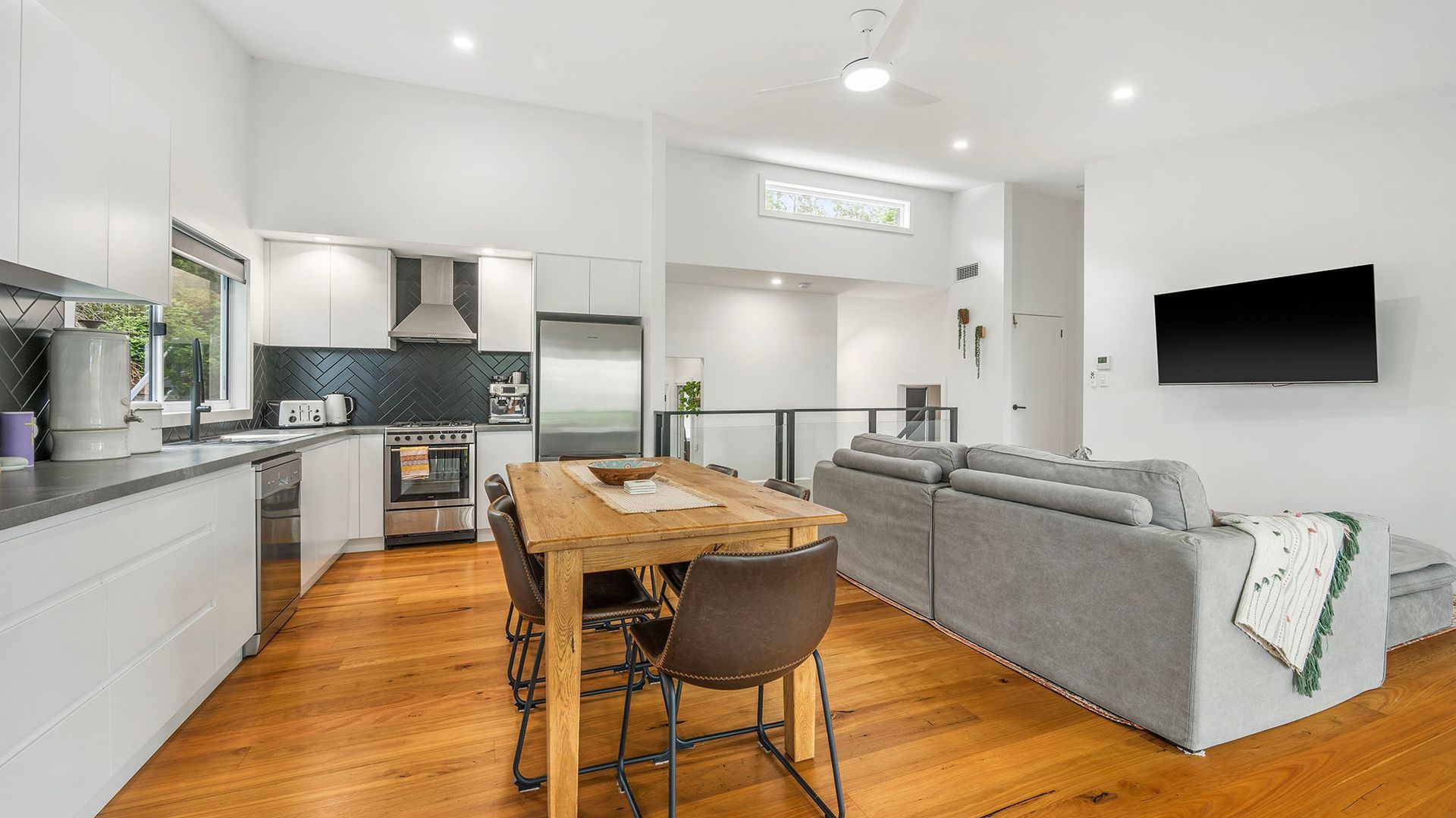 A modern kitchen with white cabinets, a wooden dining table, and a grey sofa in a living area with a flat-screen TV.