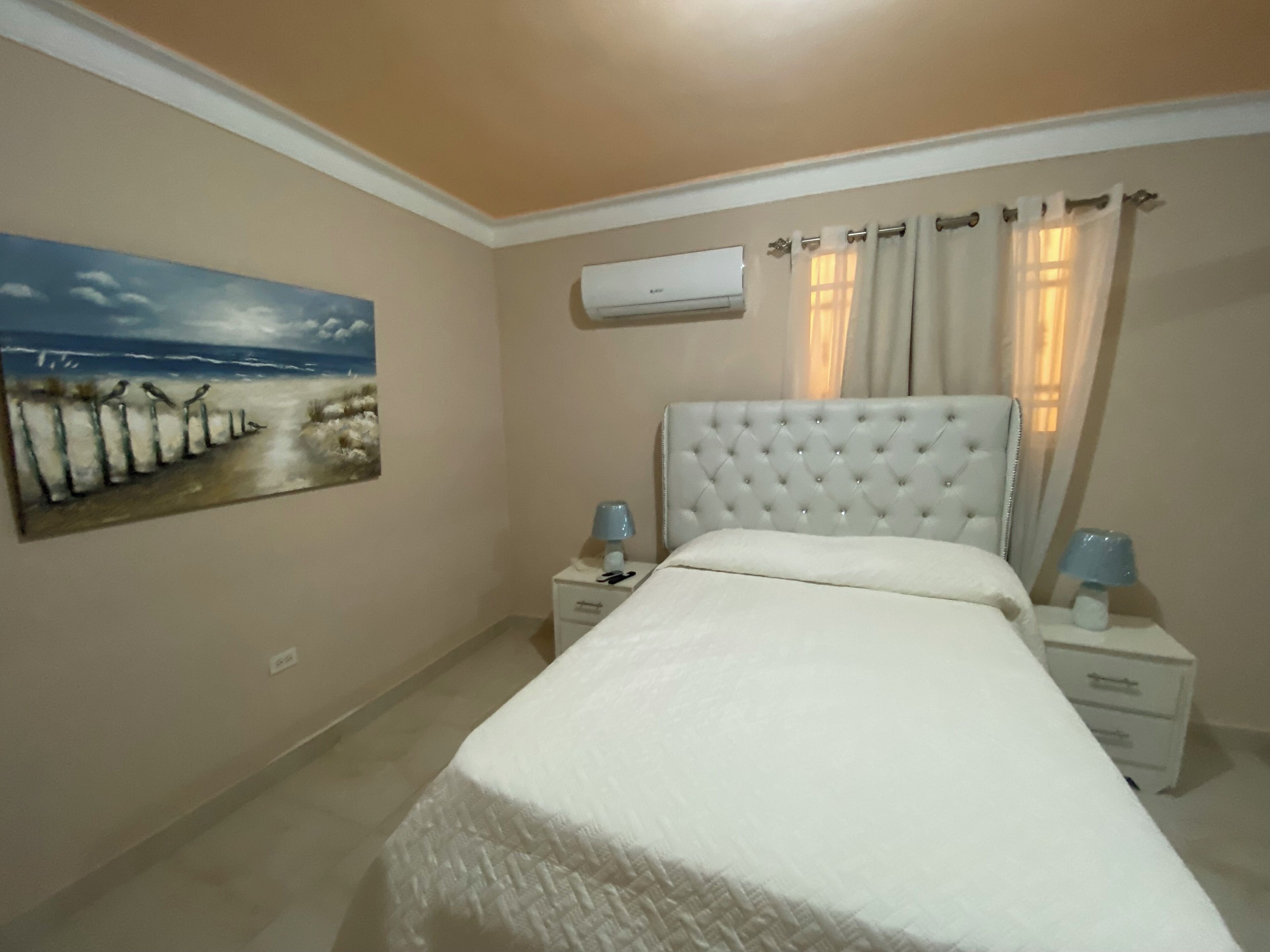 Een slaapkamer met een bed, nachtkastjes, een airconditioner en een schilderij aan de muur.