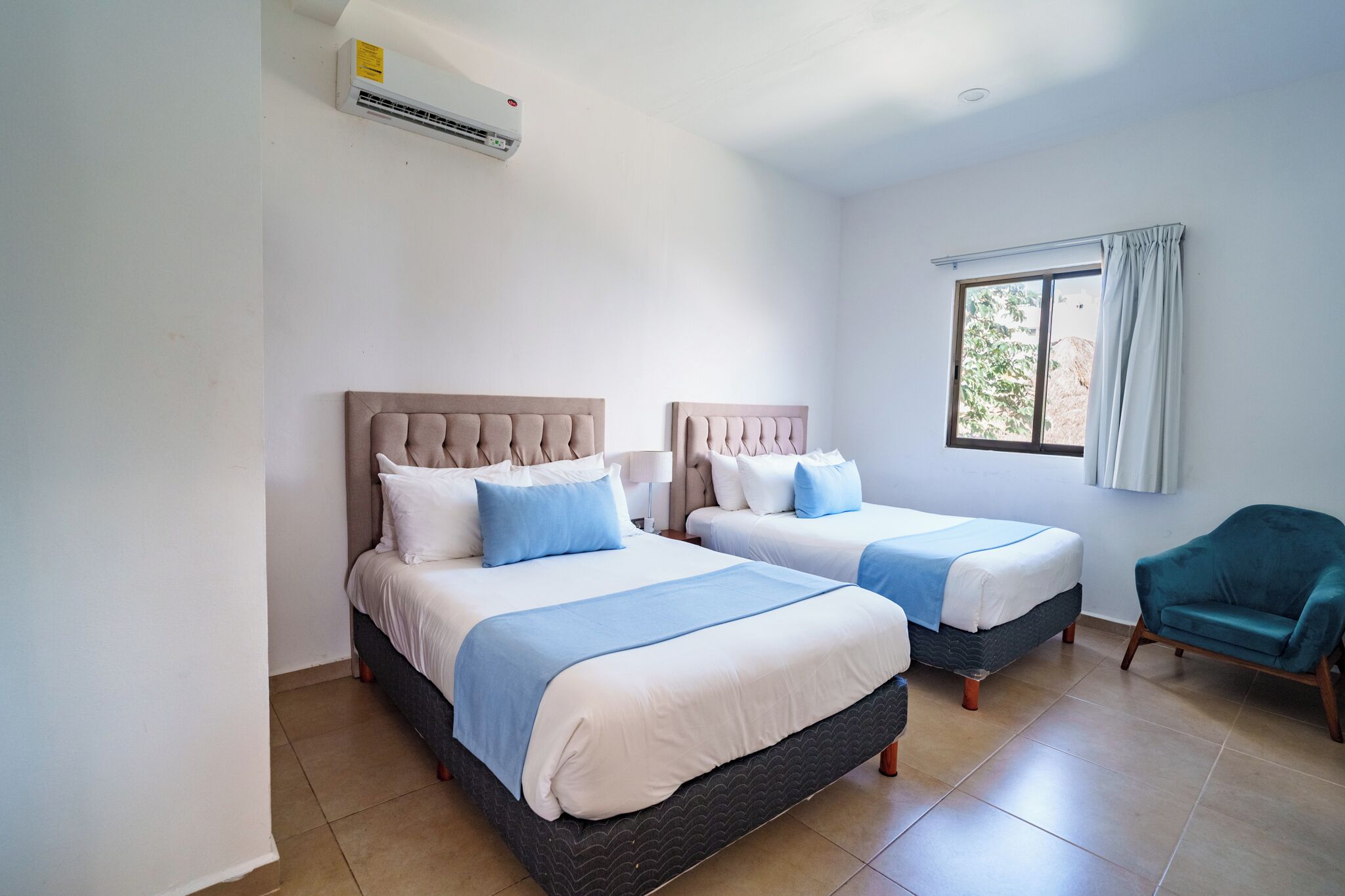 Een hotelkamer met twee bedden, een raam met gordijnen, een blauwgroene fauteuil en een airconditioning.