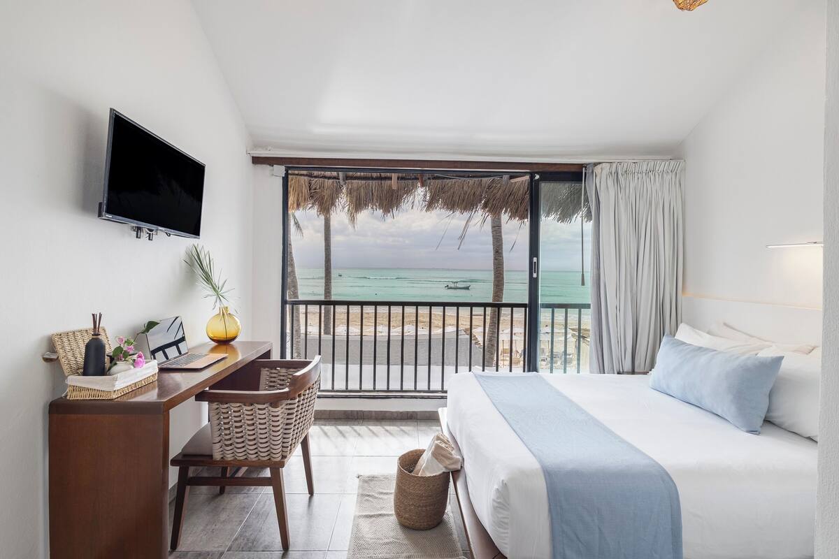 Ein Hotelzimmer mit Bett, Schreibtisch, Fernseher und Blick auf den Strand.