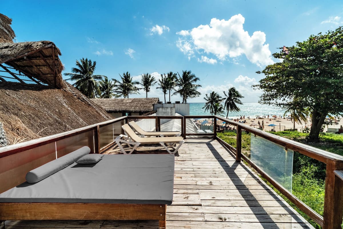 Eine Holzterrasse mit Liegestühlen und einer Liege, mit Blick auf einen Strand mit Palmen und einem klaren Himmel.