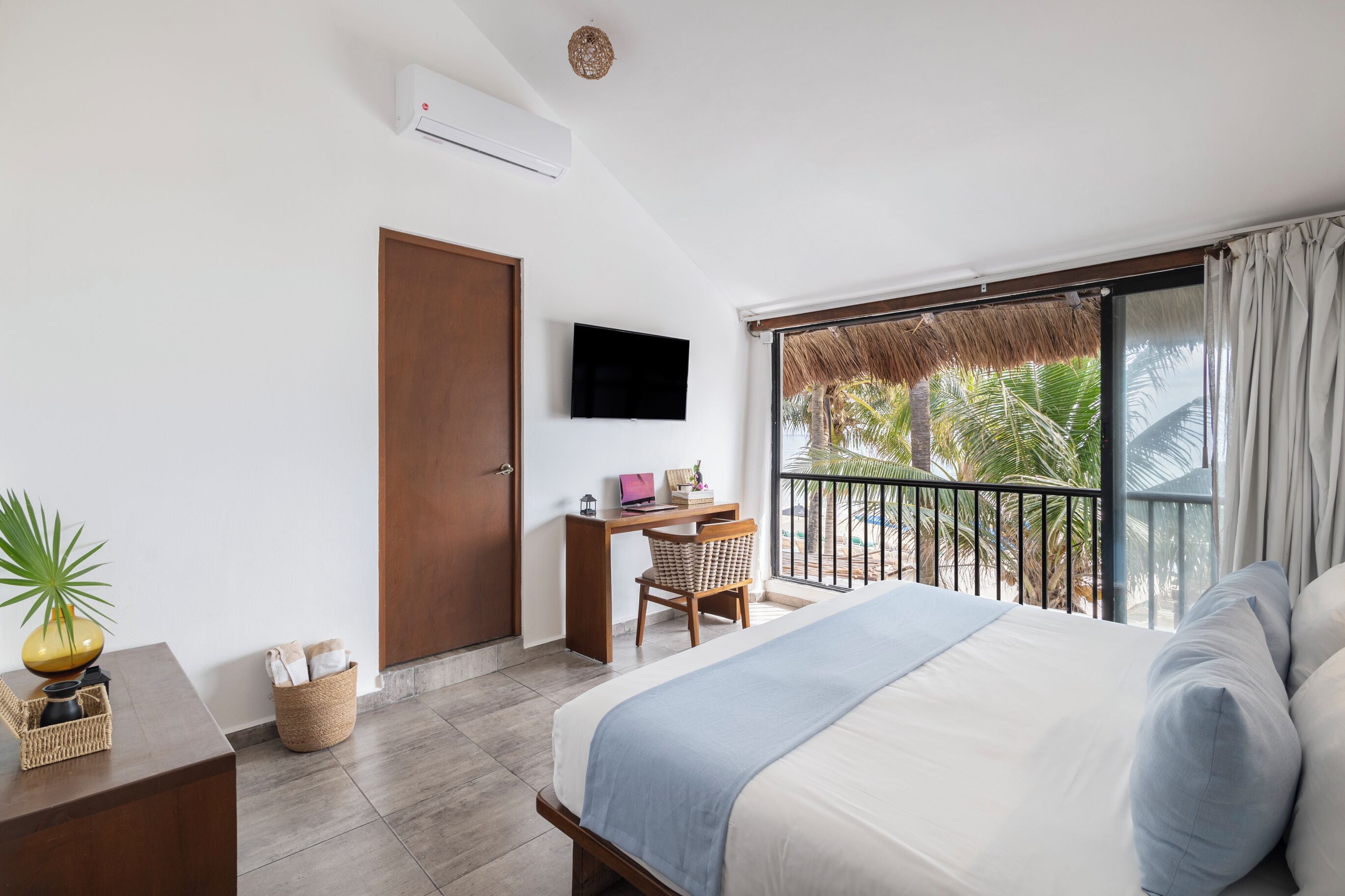 Habitación de hotel con una cama, un escritorio con silla, televisor y balcón con vista a palmeras.