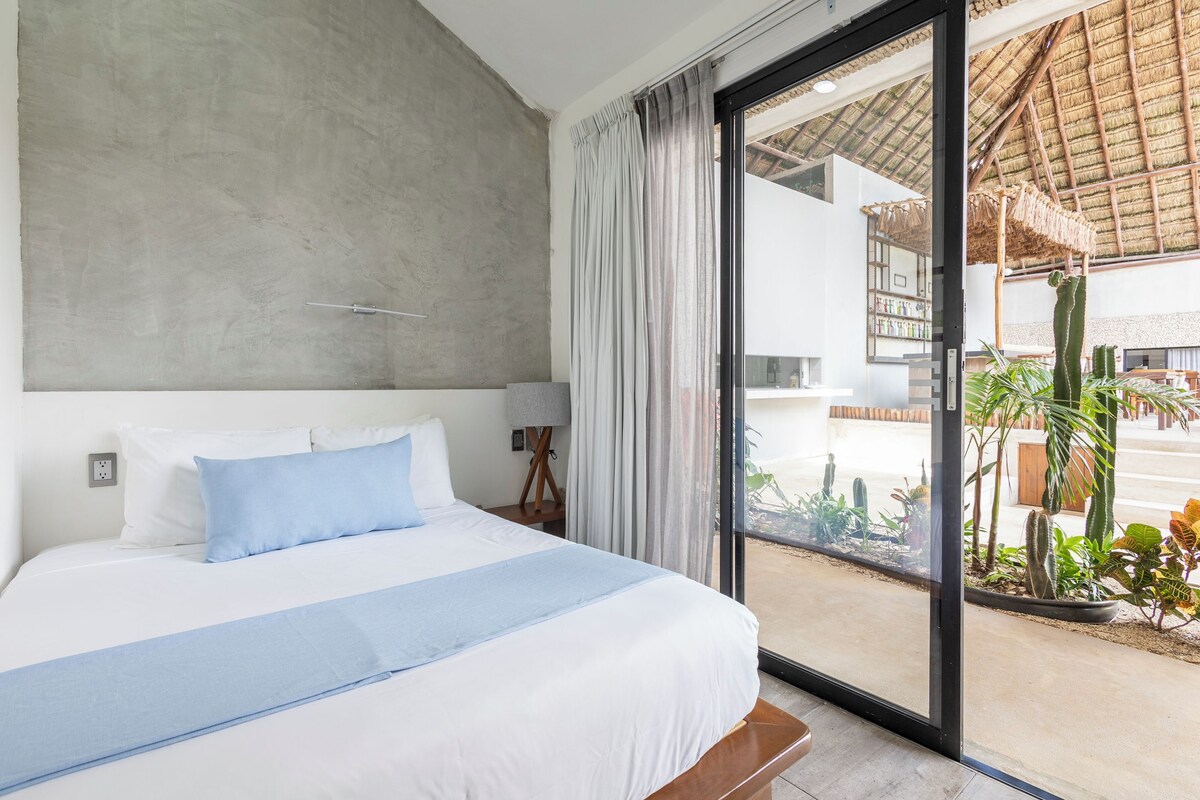 A bedroom with a large bed, a nightstand, a lamp, and a view of a patio with plants and a thatched roof.