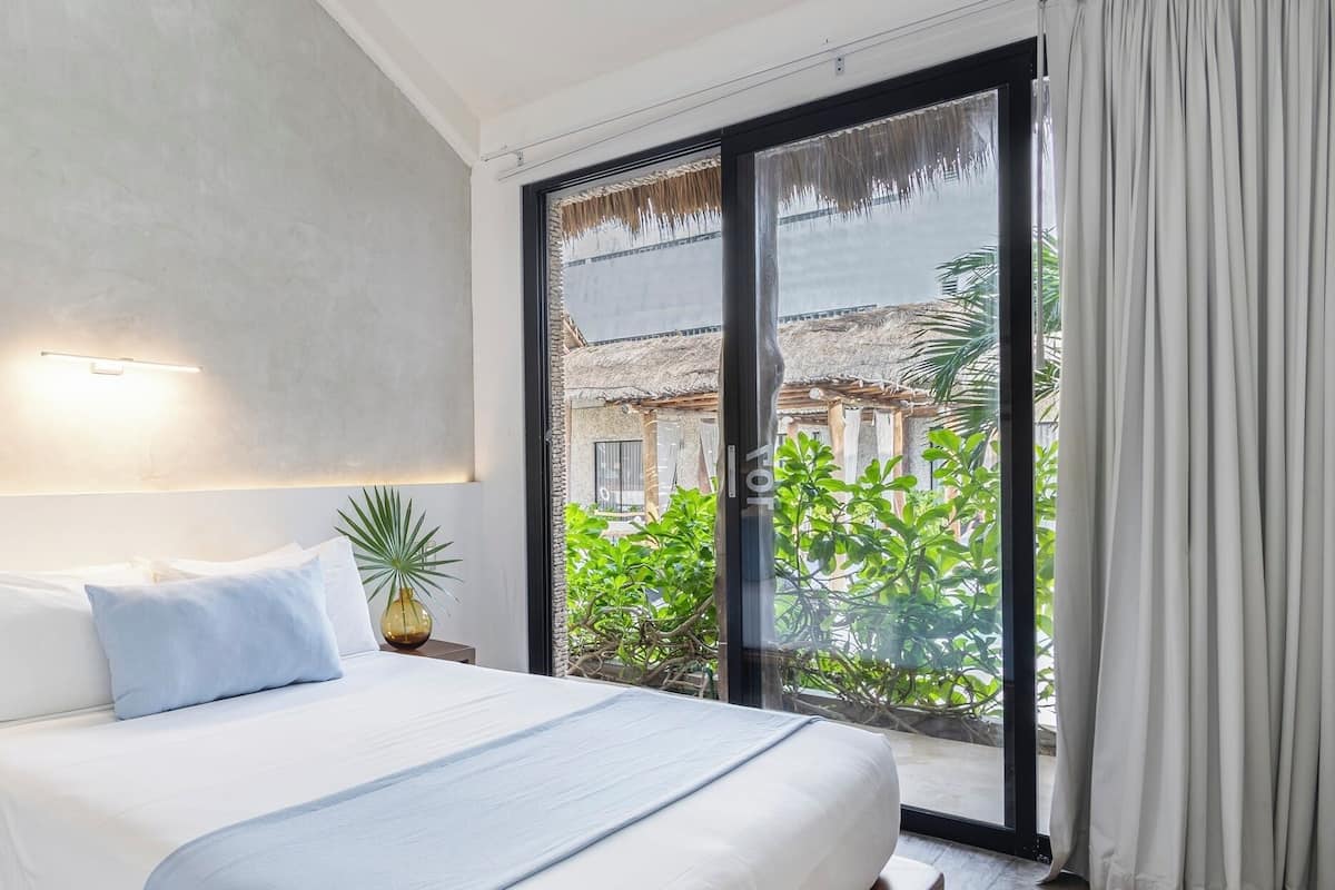 Ein modernes Hotelzimmer mit einem großen Bett, einem Nachttisch, einer Topfpflanze und Blick auf ein Gebäude mit einem Reetdach durch eine Schiebetür.