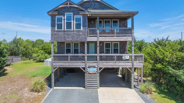 A two-story wooden house with a balcony on the second floor, a garage entrance, and a sign indicating a surfboard rental service. - Oceanside Home w/ Private Pool, Hot Tub, Game Rm. Snag this Message in a Bottle! (Salvo)