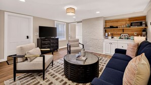 A living room with a sofa, chairs, a coffee table, a TV, a cabinet, and a kitchen area with a microwave and shelves.