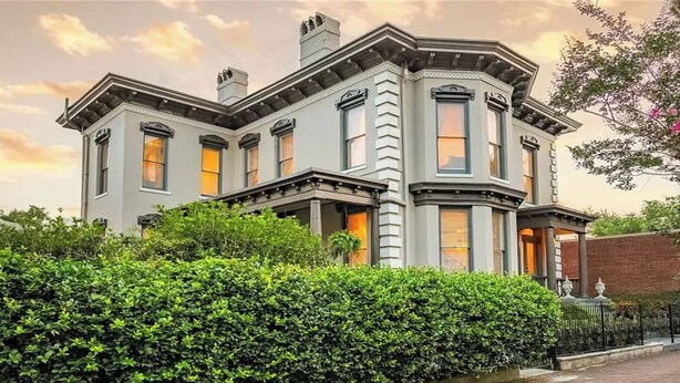 A large, historic building with a corner turret and multiple windows, surrounded by lush greenery.