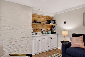 Un espace de vie compact comprenant un coin cuisine, des murs en briques blanches, un canapé bleu et une petite table d’appoint avec une lampe.