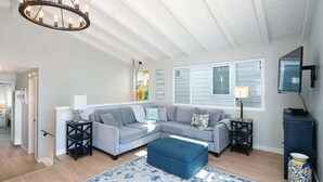 A living room with a sectional sofa, ottoman, and a chandelier. There is a TV on the wall and a window with blinds. - 304 Ruby B | Balboa Island Charmer (Newport Beach)