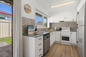 A modern kitchen with white cabinets, a built-in dishwasher, a stove, and a window overlooking a backyard. - Rose by the Bay  Coastal Getaway in Tootgarook (Tootgarook)