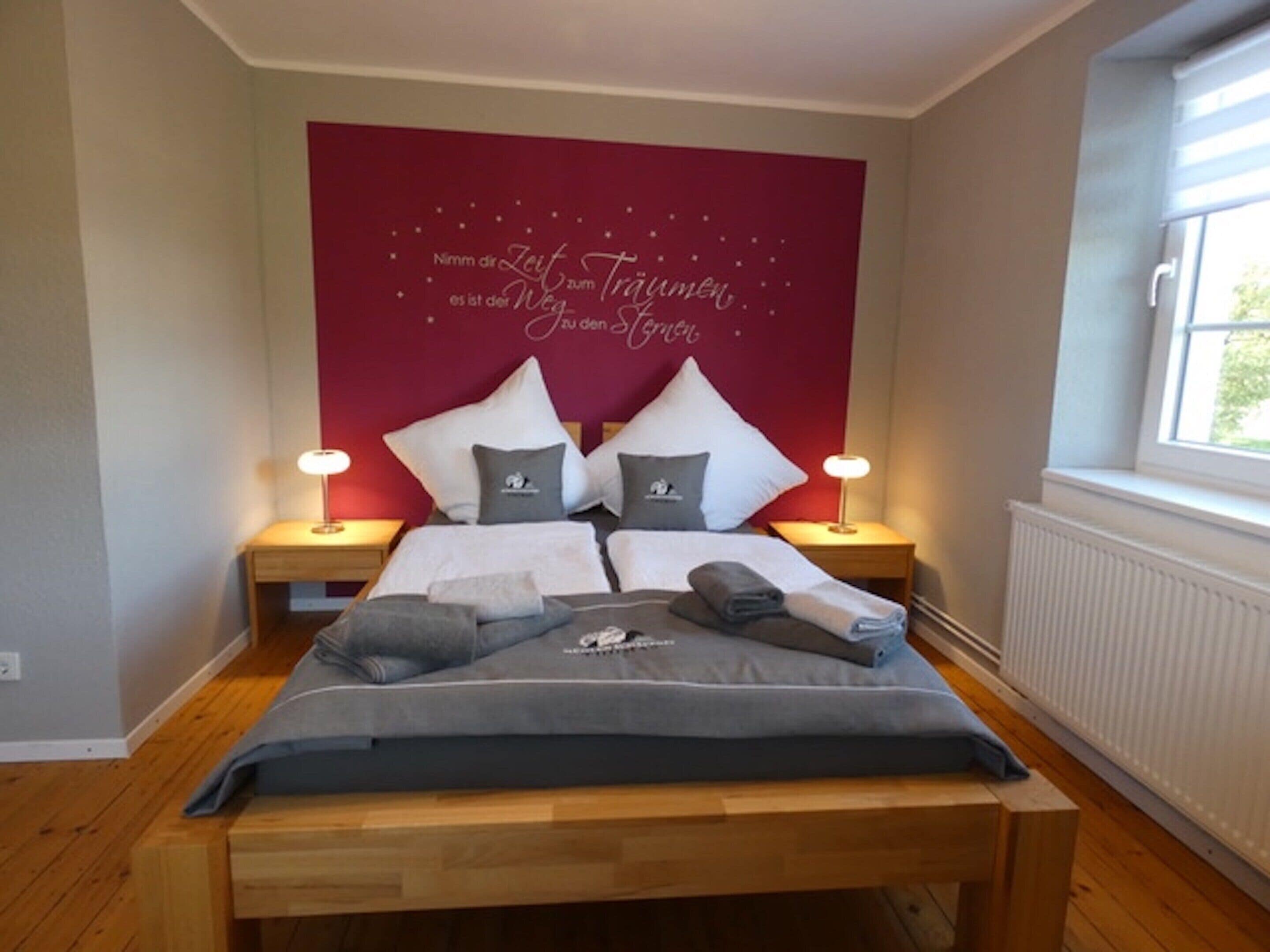 A neatly made bed with pillows and a blanket, two bedside lamps, and a red wall with a decorative phrase.
