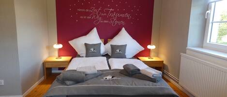 A neatly made bed with pillows and a blanket, two bedside lamps, and a red wall with a decorative phrase.