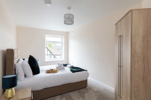 A bedroom with a bed, bedside table, lamp, wardrobe, and a window with a view of greenery. - home.ly North London Apartments Wembley (Wembley)
