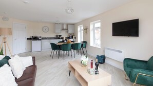 Een moderne woonkamer met een bruine bank, een houten salontafel en een groene fauteuil. Achtergrond toont een keuken met een eethoek.