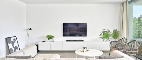 A modern living room with a white sofa, a round coffee table, and wicker chairs. A flat-screen TV is mounted on the wall above a white cabinet.