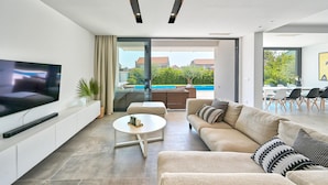 A modern living room with a flat-screen TV, a white coffee table, a beige sofa, and a glass door leading to a pool area. - 5-bedroom Villa Near Split With Pool (Kastela)