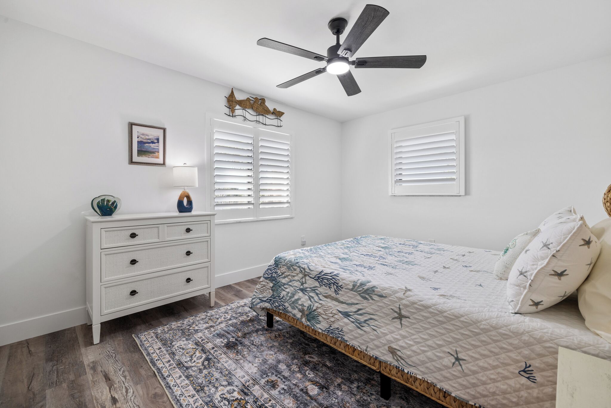 A bedroom with a bed, a ceiling fan, a nightstand, a lamp, and a window with blinds.
