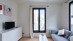 A modern living room with a white sofa, a grey sofa, a white coffee table with flowers, a TV on a white cabinet, and a large window with a view of buildings.