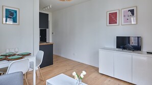 A modern living room with a dining area, a kitchen in the background, and a TV on a white cabinet.