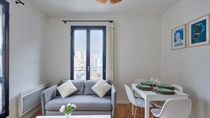 A modern living room with a grey sofa, a dining table with white chairs, and a view of a cityscape through large windows.