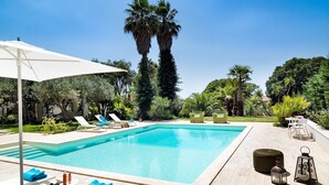 Une piscine rectangulaire, agrémentée de chaises longues et d’une table, entourée de verdure et de palmiers.