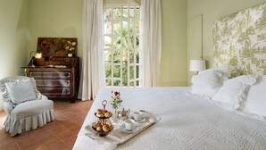 Une chambre à coucher avec un lit, une chaise, une commode, une fenêtre avec des rideaux et un plateau avec du thé et des pâtisseries.